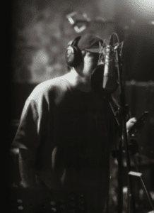 Wearing headphones and a cap, a person stands in front of a microphone in a recording studio, holding a mobile device and using SongTools to enhance their music production.